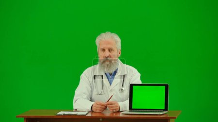 Photo for Healthcare and wellness creative concept. Portrait of man medic in studio on chroma key green screen. Senior doctor in white uniform sitting at the table with laptop with workspace mockup. Middle shot - Royalty Free Image