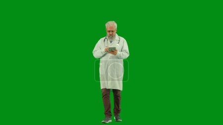 Téléchargez les photos : Santé et bien-être concept créatif. Portrait d'homme médecin en studio sur écran vert chromatique. Médecin principal en uniforme blanc marchant regardant l'information sur le comprimé. Vue de face. - en image libre de droit