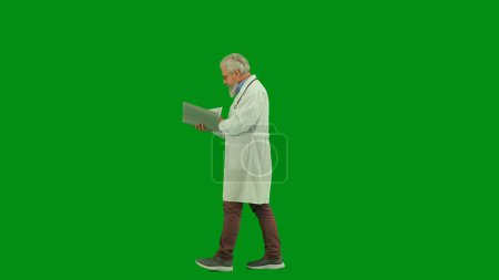 Téléchargez les photos : Santé et bien-être concept créatif. Portrait d'homme médecin en studio sur écran vert chromatique. Médecin principal en uniforme blanc marchant documents de lecture dans le dossier. Vue latérale. - en image libre de droit