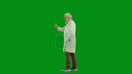 Téléchargez les photos : Santé et bien-être concept créatif. Portrait d'homme médecin en studio sur écran vert chromatique. Médecin principal en uniforme blanc marchant et saluant les gens, en agitant la main. Vue latérale. - en image libre de droit