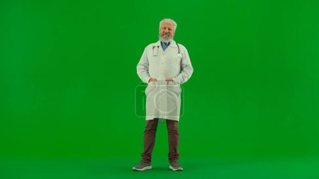 Téléchargez les photos : Santé et bien-être concept créatif. Portrait d'homme médecin en studio sur écran vert chromatique. Médecin senior en uniforme blanc debout avec les mains dans les poches et souriant. - en image libre de droit
