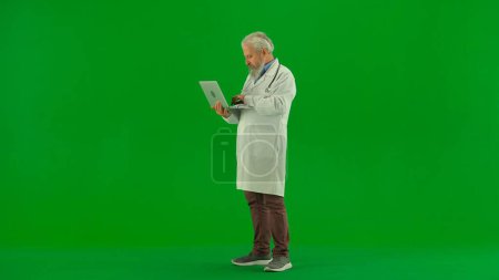 Photo for Healthcare and wellness creative concept. Portrait of man medic in studio on chroma key green screen. Senior doctor in white uniform holding laptop and reading searching for articles online. - Royalty Free Image
