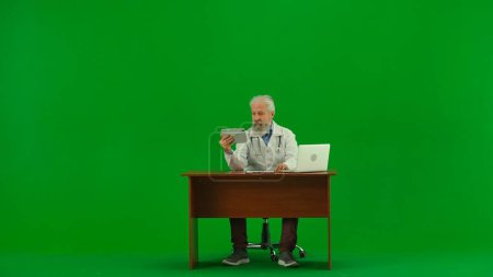 Foto de Concepto creativo de salud y bienestar. Retrato del hombre médico en el estudio en la pantalla verde croma clave. Médico senior de uniforme blanco sentado en el escritorio con portátil y mirando a la tableta. - Imagen libre de derechos