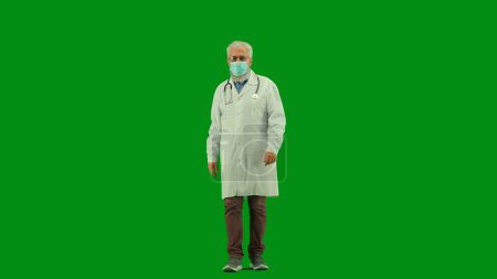 Téléchargez les photos : Santé et bien-être concept créatif. Portrait d'homme médecin en studio sur écran vert chromatique. Médecin en uniforme blanc marchant avec un masque médical. Vue de face. - en image libre de droit