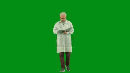 Téléchargez les photos : Santé et bien-être concept créatif. Portrait d'homme médecin en studio sur écran vert chromatique. Médecin principal en uniforme blanc marchant documents de lecture dans le dossier. Vue de face. - en image libre de droit