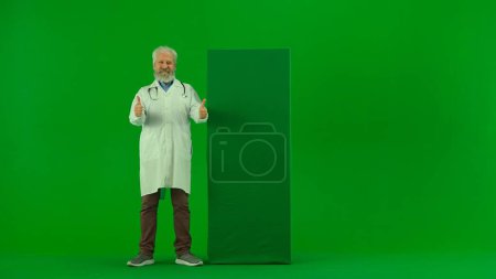 Téléchargez les photos : Santé et bien-être concept créatif. Portrait d'homme médecin en studio sur écran vert chromatique. Médecin sénior en uniforme blanc debout à côté du panneau publicitaire souriant montrant les pouces vers le haut. - en image libre de droit