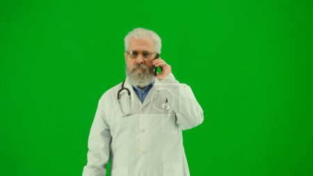 Téléchargez les photos : Santé et bien-être concept créatif. Portrait d'homme médecin en studio sur écran vert chromatique. Médecin sénior près en uniforme blanc debout et parlant sur smartphone. Plan du milieu. - en image libre de droit