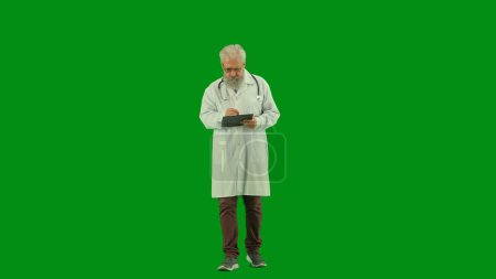 Téléchargez les photos : Santé et bien-être concept créatif. Portrait d'homme médecin en studio sur écran vert chromatique. Médecin principal en uniforme blanc tenant presse-papiers et écriture, travaillant avec des documents. Vue de face. - en image libre de droit