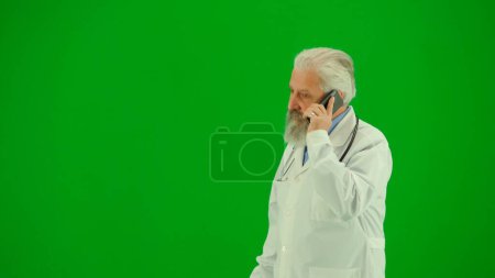 Foto de Concepto creativo de salud y bienestar. Retrato del hombre médico en el estudio en la pantalla verde croma clave. Médico senior de cerca en uniforme blanco de pie y hablando en el teléfono inteligente. Tiro del lado medio. - Imagen libre de derechos