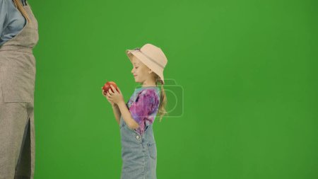 Téléchargez les photos : Jardinage et agriculture concept de publicité créative. Portrait de fille en robe et bottes en caoutchouc sur écran vert clé chroma. Petite fille mignonne jardinier tenant pomme rouge mûre dans les mains. - en image libre de droit