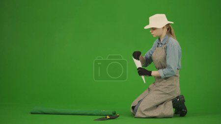 Photo for Agriculture and natural farming creative advertisement concept. Portrait of farmer in working clothing on chroma key green screen. Gardener woman sitting and planting seeds into the soil. - Royalty Free Image