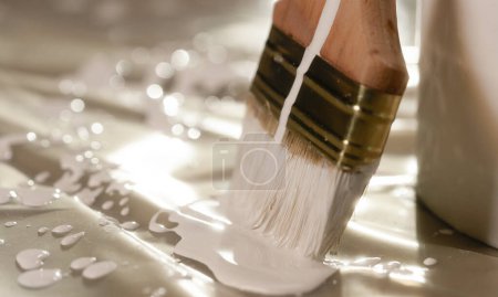 Foto de Un primer plano de un pincel con pintura blanca en su pila descansando sobre un cubo de pintura. Están de pie en un piso cubierto de tela de aceite. La pintura gotea de ella, formando un charco. - Imagen libre de derechos