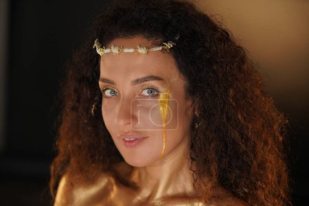 Photo for Beauty and glamour creative advertisement concept. Portrait of female model in studio. Appealing girl wearing tiara and stylish makeup covered in golden paint with golden tear looking at the camera. - Royalty Free Image