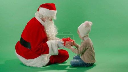 Téléchargez les photos : Santa Claude donnant boîte de cadeaux de Noël avec ruban rouge à l'enfant sur fond vert - en image libre de droit