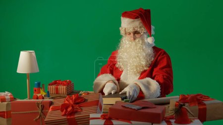Téléchargez les photos : Père Noël à la table avec des cadeaux et la lecture d'un livre avec des contes de fées sur un fond vert - en image libre de droit