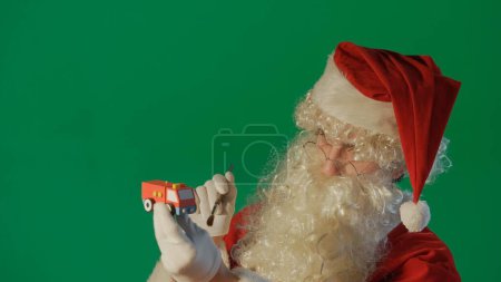 Téléchargez les photos : Portrait du Père Noël peignant une voiture avec un cadeau sur un fond vert - en image libre de droit