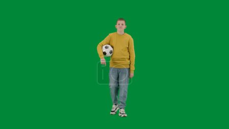 Photo for School kids and leisure time creative concept. Portrait of kid boy on chroma key green screen. Schoolboy in jeans walking holding football ball and looking around. Full body front shot. - Royalty Free Image