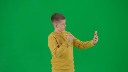 Photo for School kids and leisure time creative concept. Portrait of kid boy on chroma key green screen. Schoolboy in jeans taking selfie on smartphone showing thumbs up. Middle body side shot. - Royalty Free Image