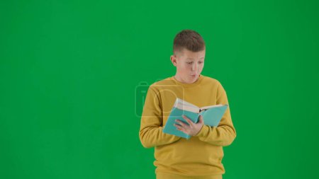 Téléchargez les photos : Les écoliers et les loisirs concept créatif. Portrait de gamin sur écran vert chromatique. Écolier en jean tenant le livre et le lisant avec une expression de visage étonnante. Plan avant du milieu du corps. - en image libre de droit