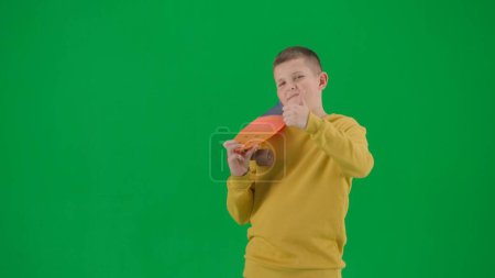 Photo for School kids and leisure time creative concept. Portrait of kid boy on chroma key green screen. Schoolboy in jeans holding skateboard looking at the camera showing thumbs up. Middle body front shot. - Royalty Free Image