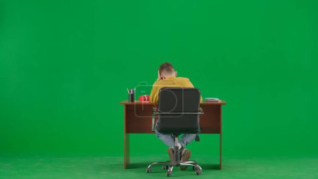 Photo for School kids and leisure time creative concept. Portrait of kid boy on chroma key green screen. Schoolboy in jeans sitting at the desk doing homework, writing in notebook, reading. Back full shot. - Royalty Free Image
