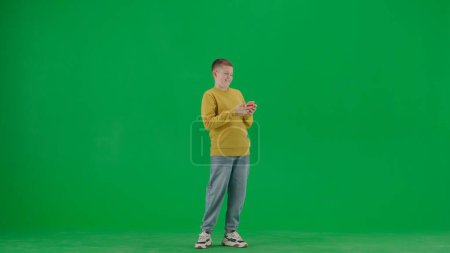 Photo for School kids and leisure time creative concept. Portrait of kid boy on chroma key green screen. Schoolboy in jeans standing watching social media funny videos on smartphone. Full body shot. - Royalty Free Image