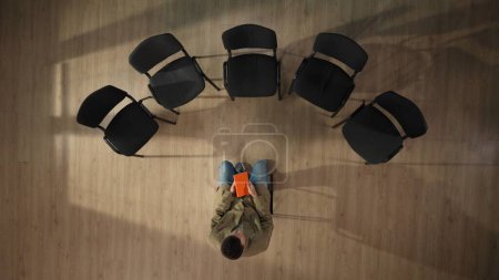 Foto de En el marco de arriba, un hombre de mediana edad está sentado en una silla en el centro. Él está esperando que su reunión de grupo comience, pensando en algo, pensando. Está sosteniendo un cuaderno naranja. Vista superior. - Imagen libre de derechos