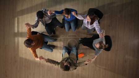 Photo for In the shot above a group of people, a man and a woman and their mentor are sitting on a chair. They have come to a meeting, a session, therapy. They are sitting in a circle holding hands. Top view. - Royalty Free Image