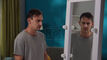 Foto de Hombre agotado mirando su reflejo en el espejo, de pie en la sala de estar de cerca. Apatía, conflicto interno, problemas psicológicos - Imagen libre de derechos