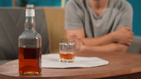 Téléchargez les photos : Une bouteille de whisky et un support en verre sur la table se referment. L'alcool se dresse sur un fond flou d'un homme assis sur un canapé et se serrant dans ses bras. Alcoolisme et troubles mentaux - en image libre de droit