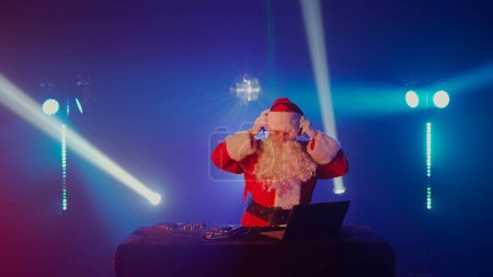 Foto de DJ Santa Claus mezclando pistas en un club nocturno en una fiesta de Navidad y Año Nuevo o eventos corporativos. Senior disc jockey como Santa escuchar música, auriculares, ordenador portátil, reproductor de controlador de mezclador, tocadiscos - Imagen libre de derechos