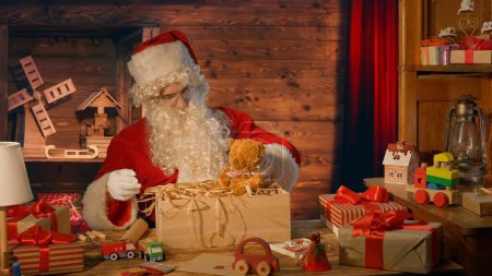 Foto de Santa Claus en la mesa con regalos y mirando a un oso de peluche en su casa - Imagen libre de derechos