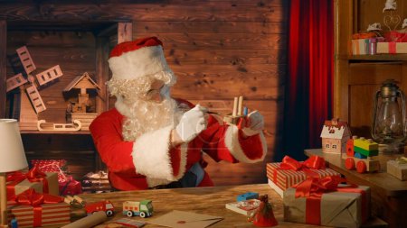 Photo for Santa Claus at the table with gifts and paints a wooden toy at his house - Royalty Free Image