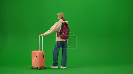 Foto de Concepto de publicidad creativa del aeropuerto. Retrato de persona turística aislado en croma pantalla verde clave de fondo. Mujer joven con la maleta de pie y mirando el tablero de información. - Imagen libre de derechos