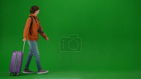 Photo for Airport creative advertisement concept. Portrait of person tourist isolated on chroma key green screen background. Young man with suitcase walking with suitcase in orange hoodie and jeans. - Royalty Free Image