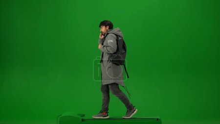 Photo for Airport creative advertisement concept. Portrait of person tourist isolated on chroma key green screen background. Adult man in winter coat with backpack walking and talking on smartphone. - Royalty Free Image