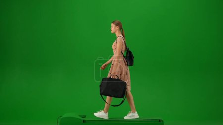 Foto de Concepto de publicidad creativa del aeropuerto. Retrato de persona turística aislado en croma pantalla verde clave de fondo. Mujer joven con bolsa para portátil en vestido caminando y mirando a su alrededor. - Imagen libre de derechos