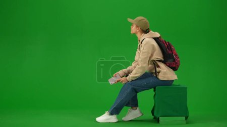 Foto de Concepto de publicidad creativa del aeropuerto. Retrato de persona turística aislado en croma pantalla verde clave de fondo. Mujer joven sentada sosteniendo un teléfono inteligente mirando el tablero de información. - Imagen libre de derechos