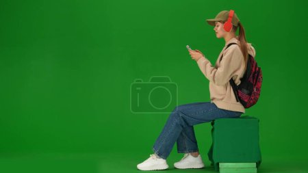 Téléchargez les photos : Concept publicitaire créatif aéroport. Portrait de touriste de personne isolé sur fond d'écran vert clé chroma. Jeune femme assise tenant smartphone écoutant de la musique dans un casque. - en image libre de droit