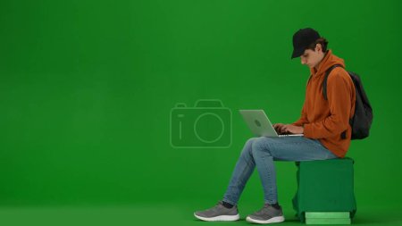 Photo for Airport creative advertisement concept. Portrait of person tourist isolated on chroma key green screen background. Young man sitting holding laptop and working, waiting for flight. - Royalty Free Image