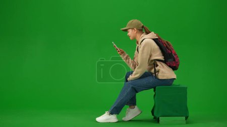Foto de Concepto de publicidad creativa del aeropuerto. Retrato de persona turística aislado en croma pantalla verde clave de fondo. Mujer joven sentada sosteniendo el teléfono inteligente mirando la pantalla expresión sonriente. - Imagen libre de derechos