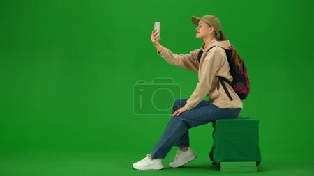 Téléchargez les photos : Concept publicitaire créatif aéroport. Portrait de touriste de personne isolé sur fond d'écran vert clé chroma. Jeune femme assise tenant smartphone et prenant des photos selfie. - en image libre de droit