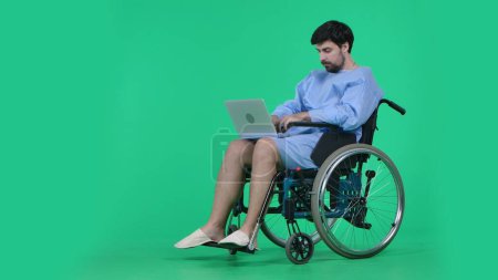 Photo for Medical ward and rehabilitation creative concept on chroma key green screen. Adult man patient in robe sitting in wheelchair holding laptop and typing, working online, focused face expression. - Royalty Free Image