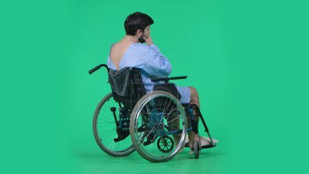 Photo for Medical ward and rehabilitation creative concept on chroma key green screen. Adult man patient in robe sitting in wheelchair shot from a back, sad face expression, holding head in hand. - Royalty Free Image