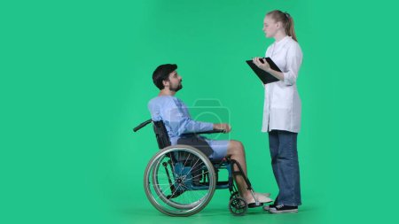 Photo for Medical ward and rehabilitation creative concept on chroma key green screen. Adult man patient in robe sitting in wheelchair, female nurse with clipboard standing next to him asking questions. - Royalty Free Image