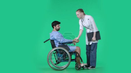 Téléchargez les photos : Service médical et concept créatif de réhabilitation sur écran vert chromatique. Homme adulte patient en peignoir assis en fauteuil roulant, infirmière debout à côté de lui vérifiant pouls homme. - en image libre de droit