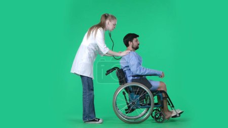 Photo for Medical ward and rehabilitation creative concept on chroma key green screen. Adult man patient in robe sitting in wheelchair, female nurse with stethoscope checking his lungs breathing on the back. - Royalty Free Image