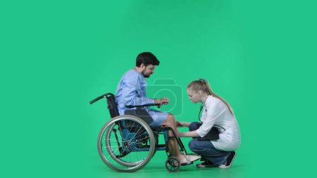 Photo for Medical ward and rehabilitation creative concept on chroma key green screen. Adult man patient in robe sitting in wheelchair, female nurse checking his legs, touching feet. - Royalty Free Image