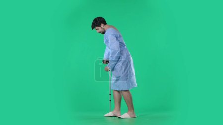 Photo for Medical ward and rehabilitation creative concept on chroma key green screen. Adult man patient in hospital robe holding crutch and making few successful steps with support. - Royalty Free Image