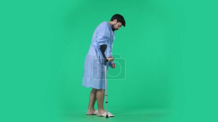 Photo for Medical ward and rehabilitation creative concept on chroma key green screen. Adult man patient in hospital robe holding crutch and making few successful steps, walking with support. - Royalty Free Image